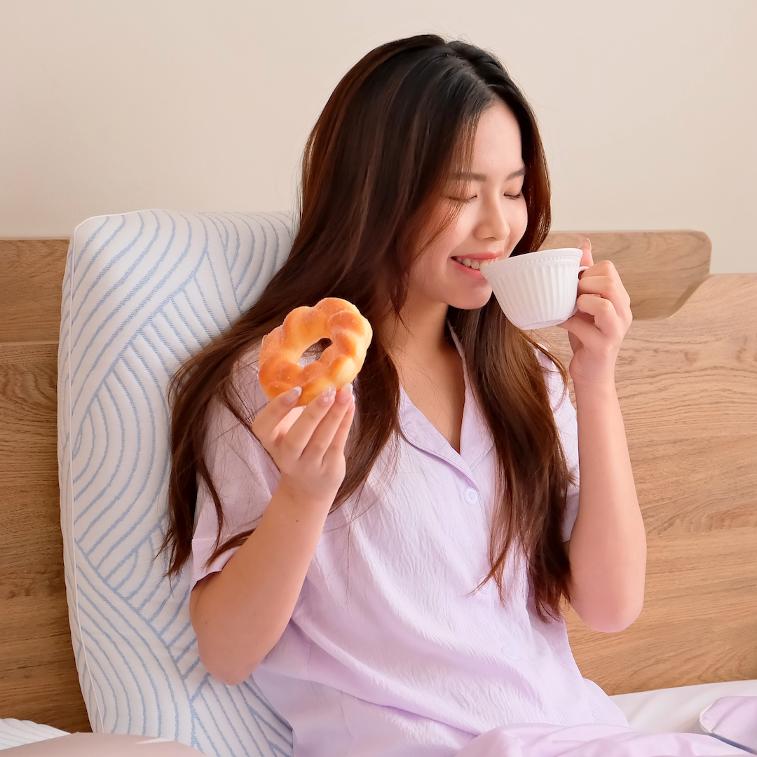 Woman in pajamas holding a donut and a cup leaning on Nemuri Pillow on a bed